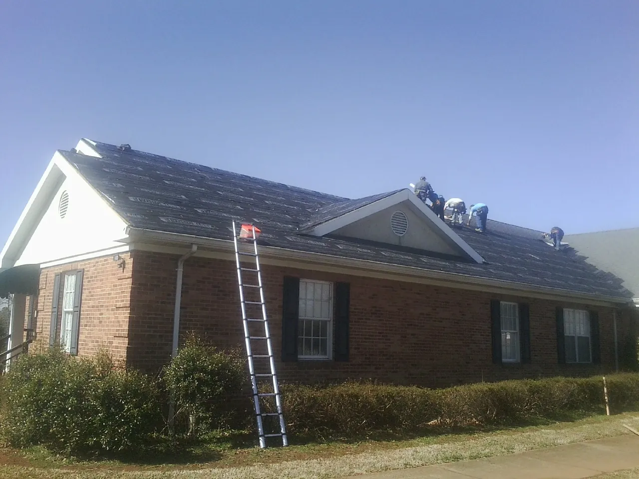 Crew tearing shingles off a brick ranch roof with a ladder set — Emergency Roof Repair work by our crew in Urbandale