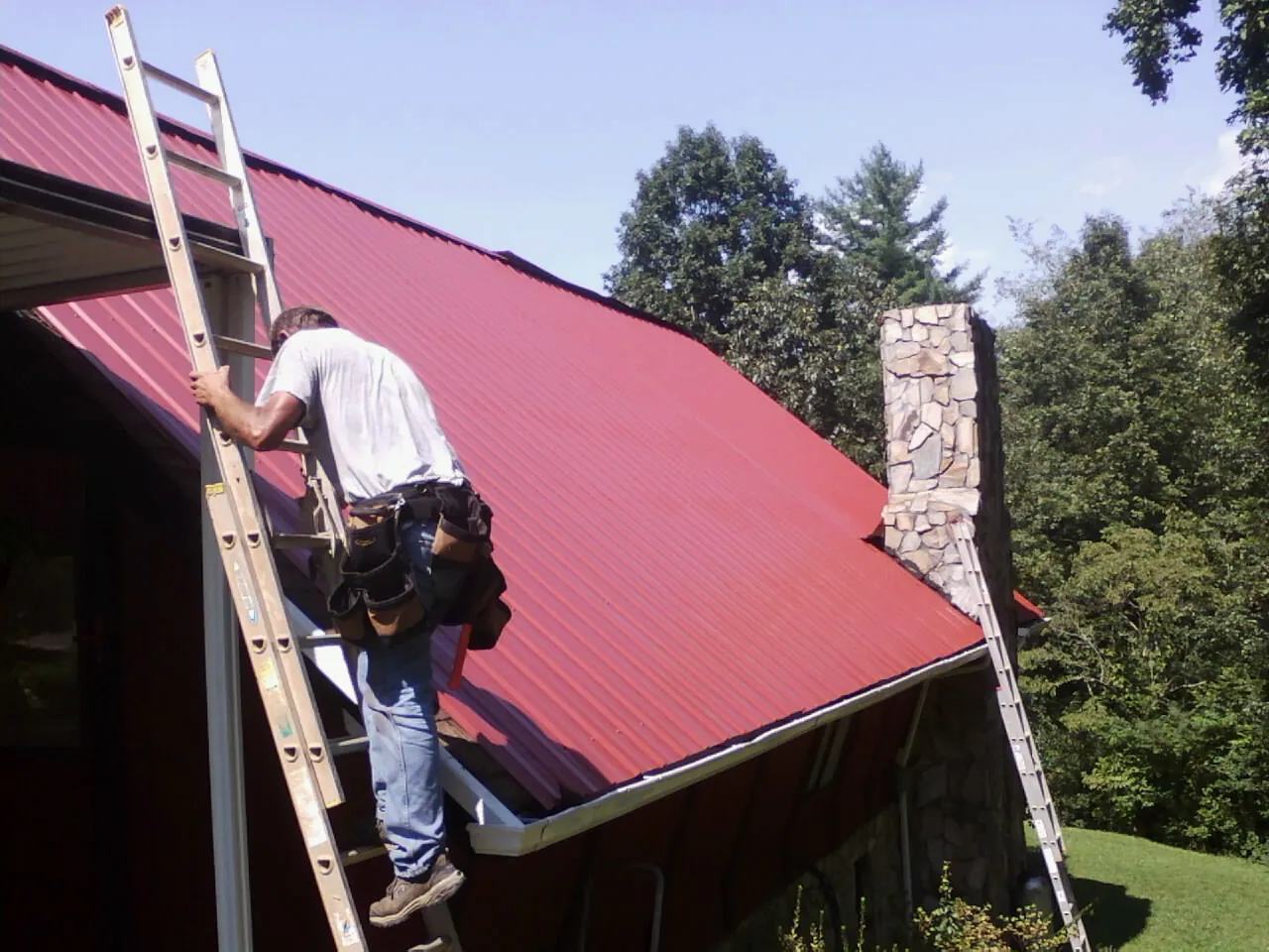 Roofer on a ladder beside a red metal roof and stone chimney — Soffit & Fascia Repair by Roofing Brothers in Urbandale